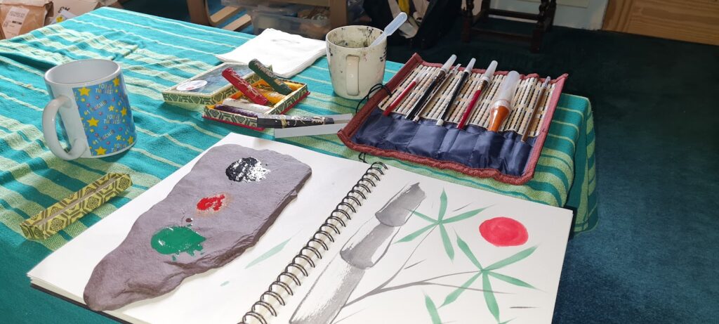 Image shows a partically completed drawing of a bamboo plant in a sketchbook. Near this is a set of chinese brush pens, stick inks and a slate inkstone.