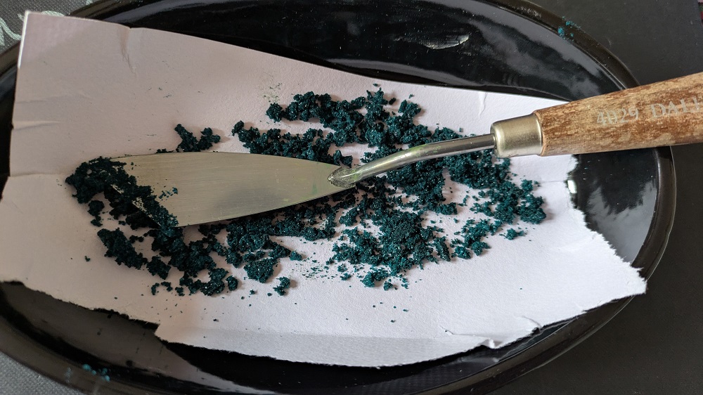 A black dish containing blotting paper, a pallate knife and green crystallline precipitate